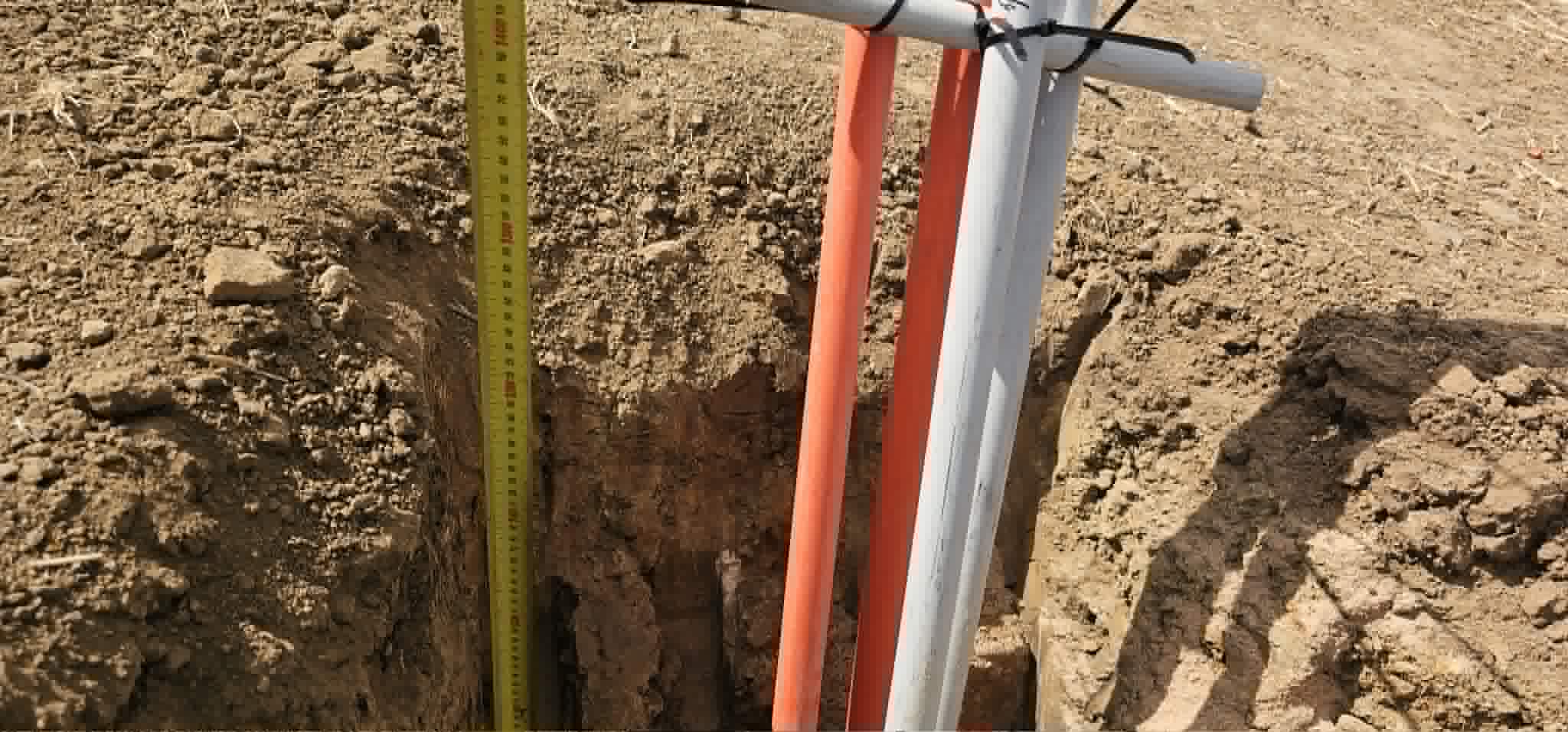 Tape measure checking trench depth beside orange power and white communications conduits