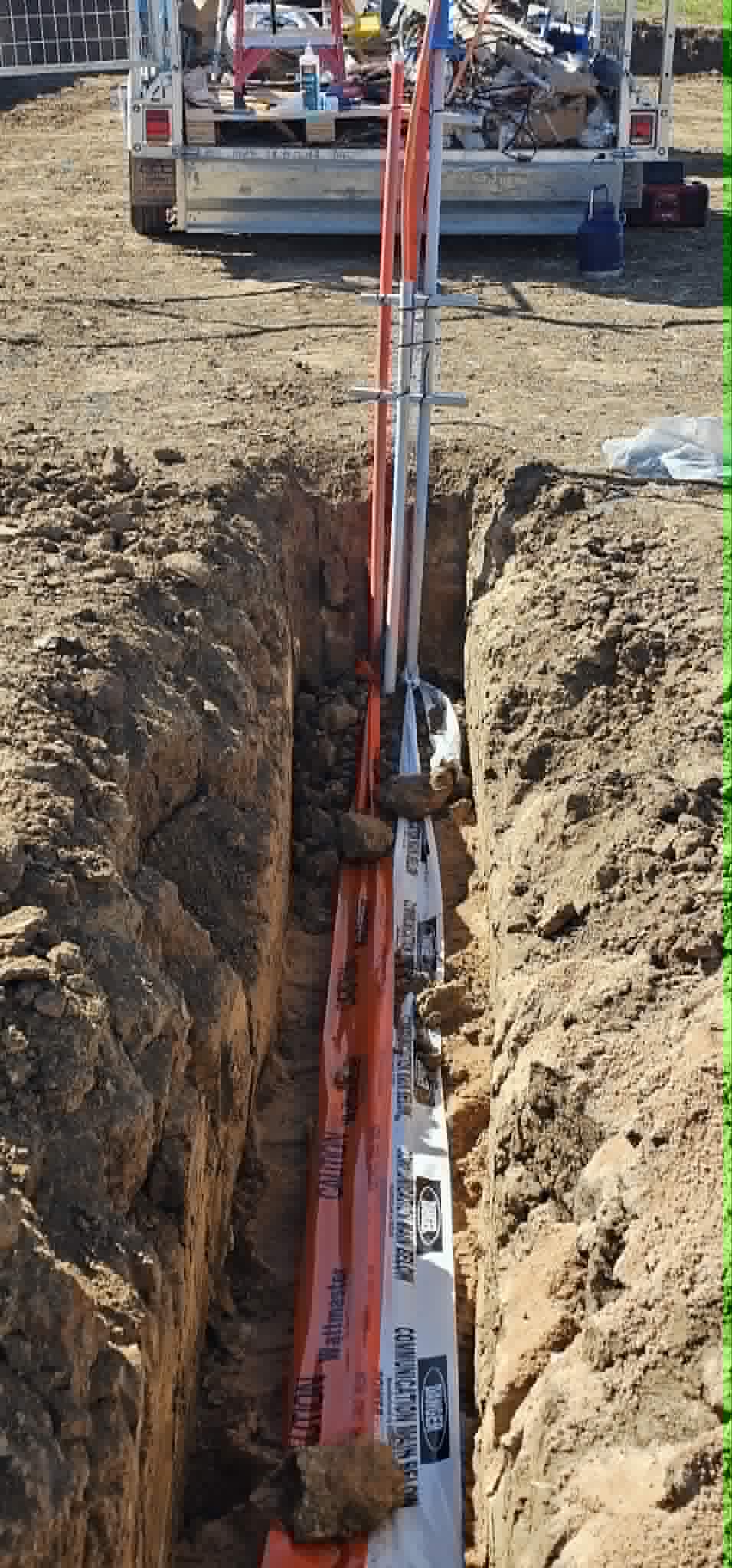 Open trench with orange power conduit and white communications conduit running to a trailer